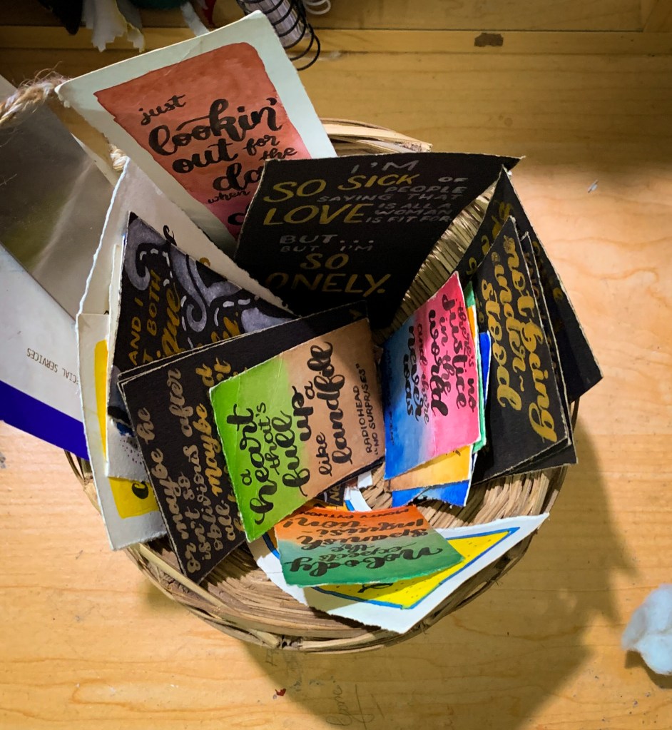 A photo of a basket filled with paper bookmarks of various colours and sizes, each bearing quotes from books or song lyrics.